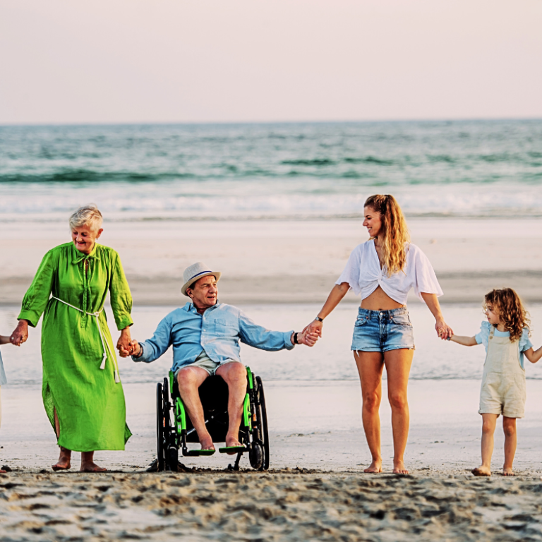 A family holding hands on the beach at sunset, including a man in a wheelchair.